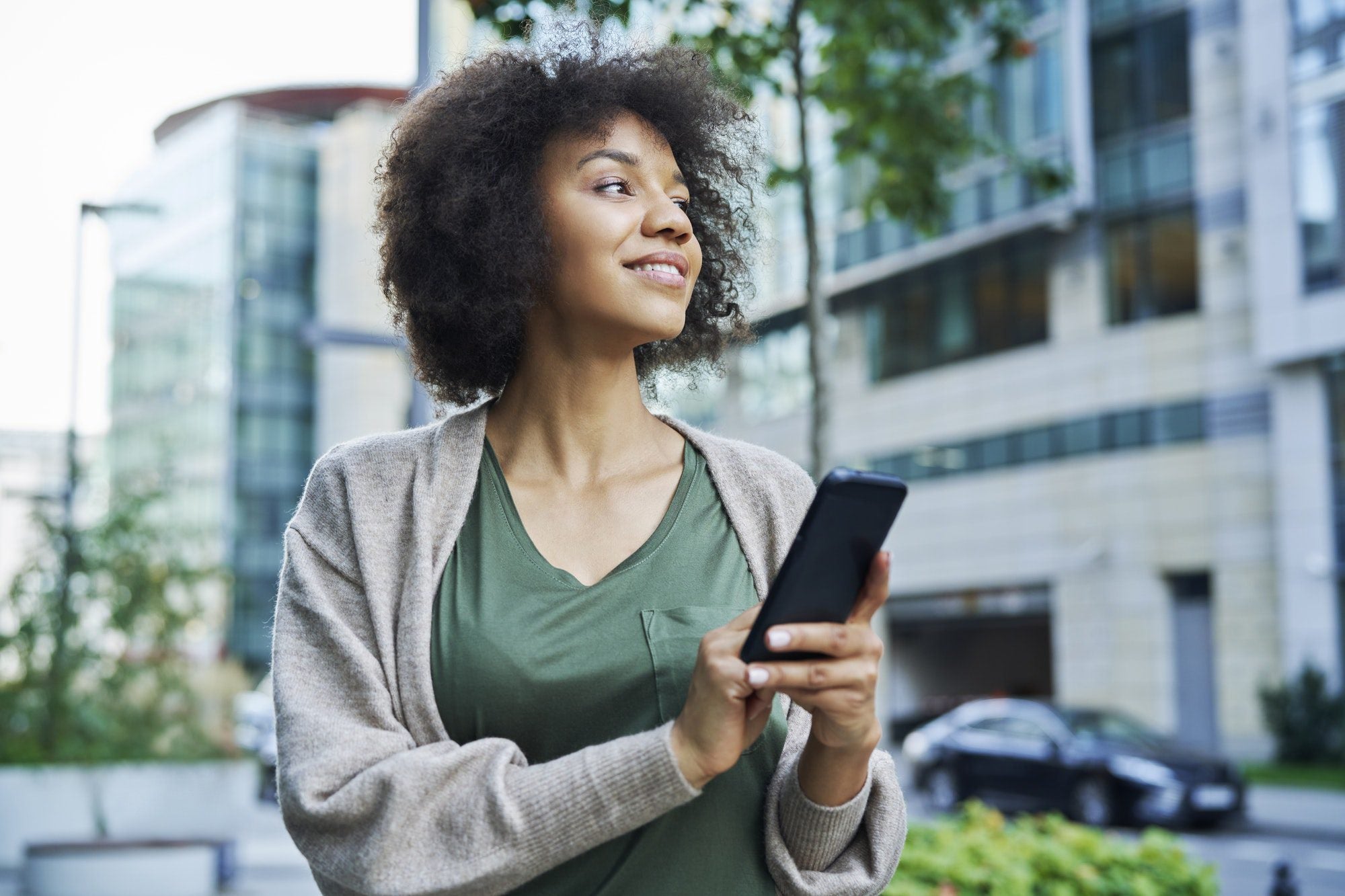 A woman outdoors, holding a tablet in front of her. She is dressed in casual street fashion, with a building and a car visible in the background.