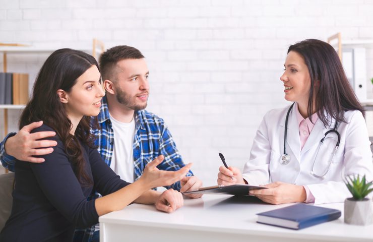 A small group of people gathered indoors around a table, engaged in a discussion. They are smiling and collaborating, with various clothing styles visible. The setting appears to be a classroom or meeting room, with a wall in the background.